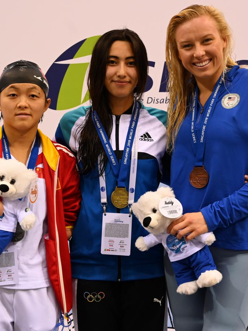 three female swimmers on the podium holding their medals and mascot teddys
