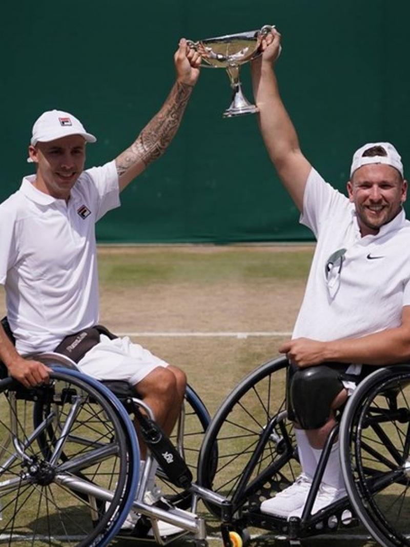 Two male wheelchair tennis players holding a trophy on a grass court