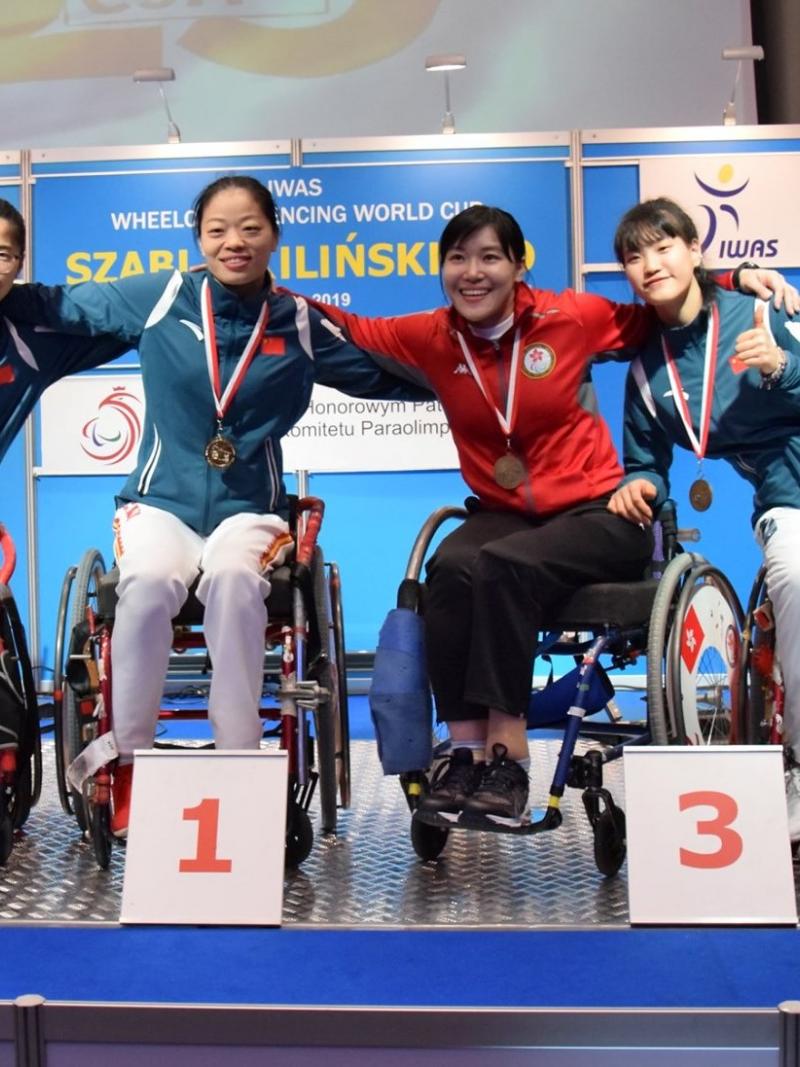 Four female wheelchair fencers put arms around each other and pose on a podium