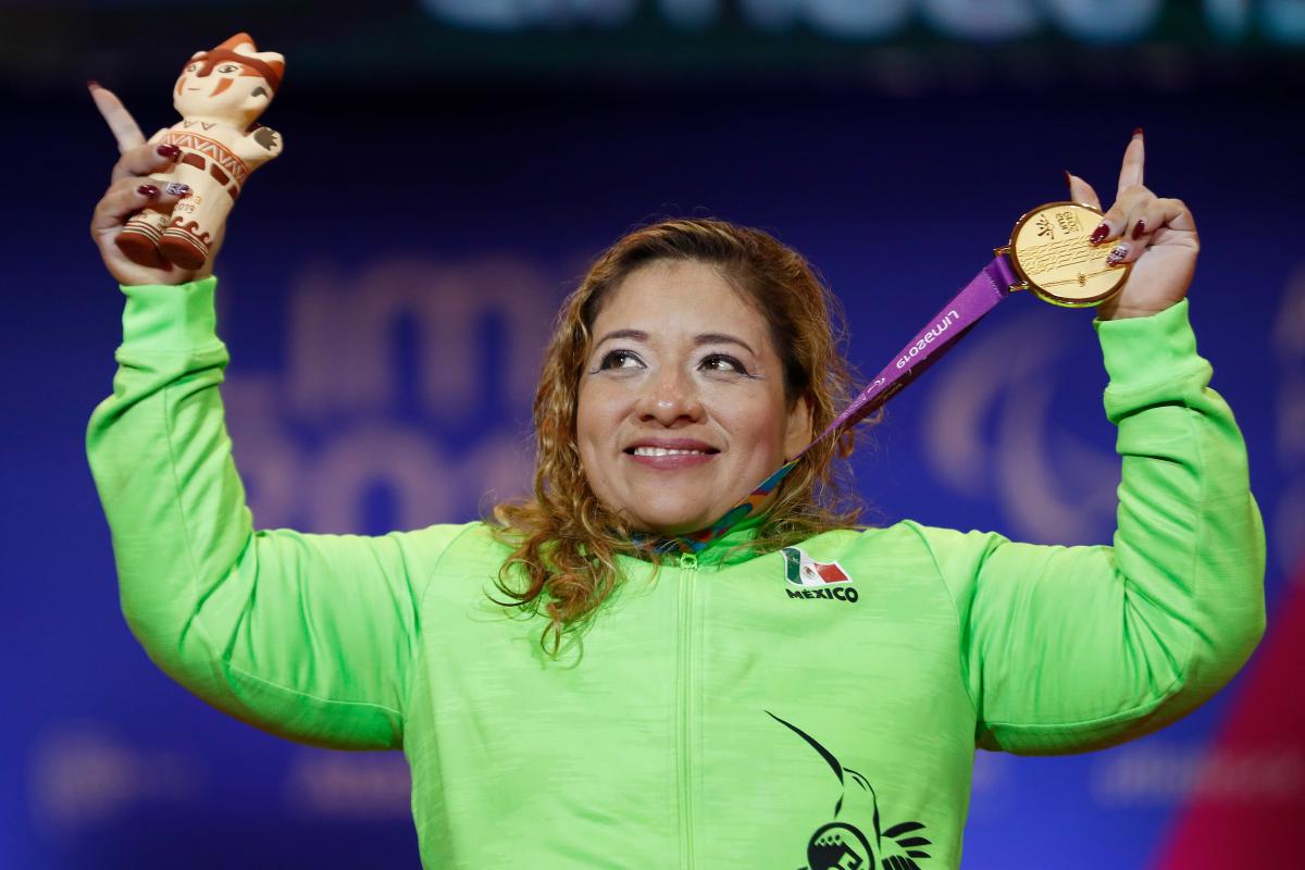 a female powerlifter holds up her gold medal and points her fingers to the sky