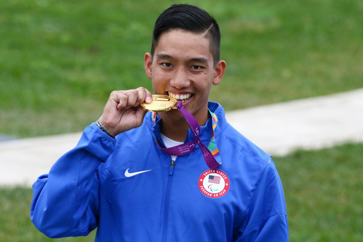 a male Para shooter biting a gold medal and smiling
