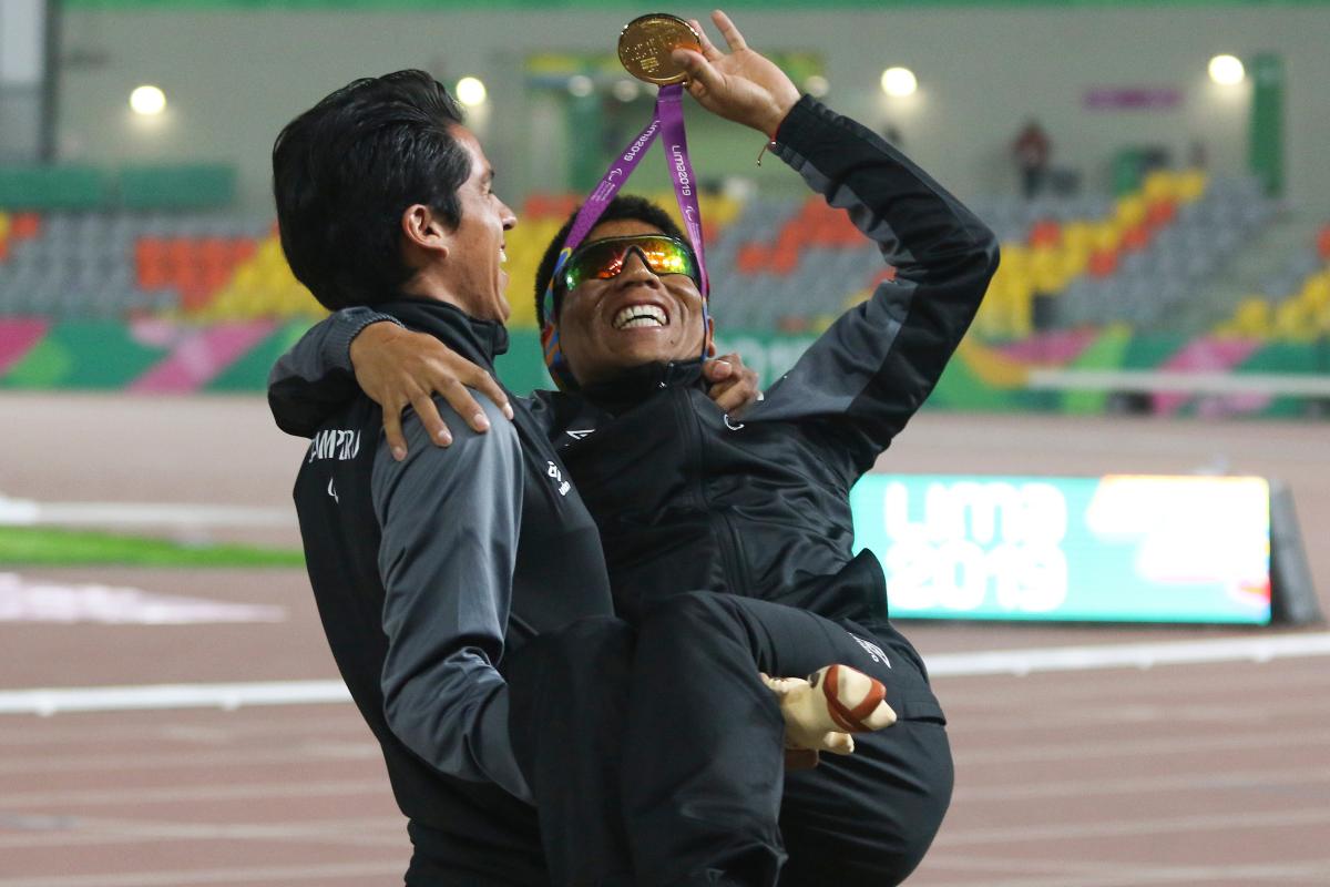 a male vision impaired runner being carried by his guide while waving his medal