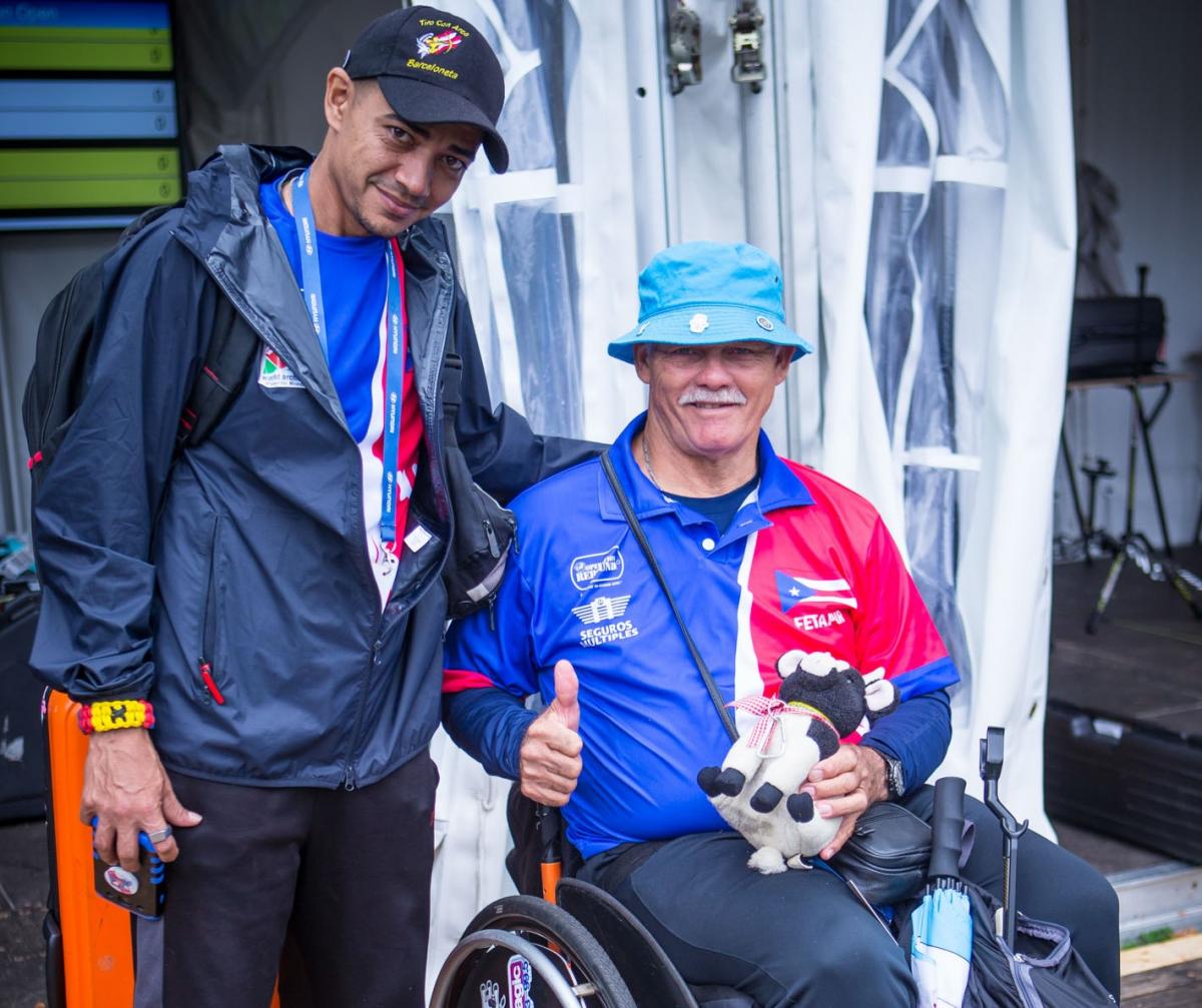 two men, one standing, one sitting in a wheelchair giving a thumbs up