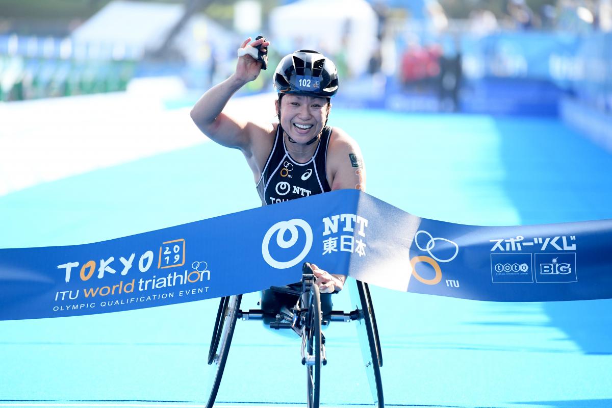a female Para triathlete in a wheelchair raises her arm as she breaks the finish line tape