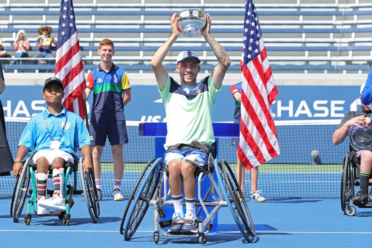 a male wheelchair tennis player holds up a trophy