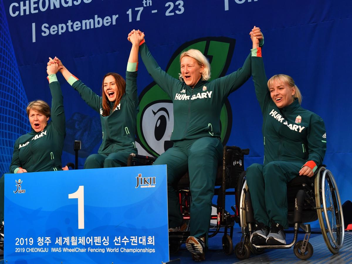 Four Hungarian female wheelchair fencers hold hands on the podium