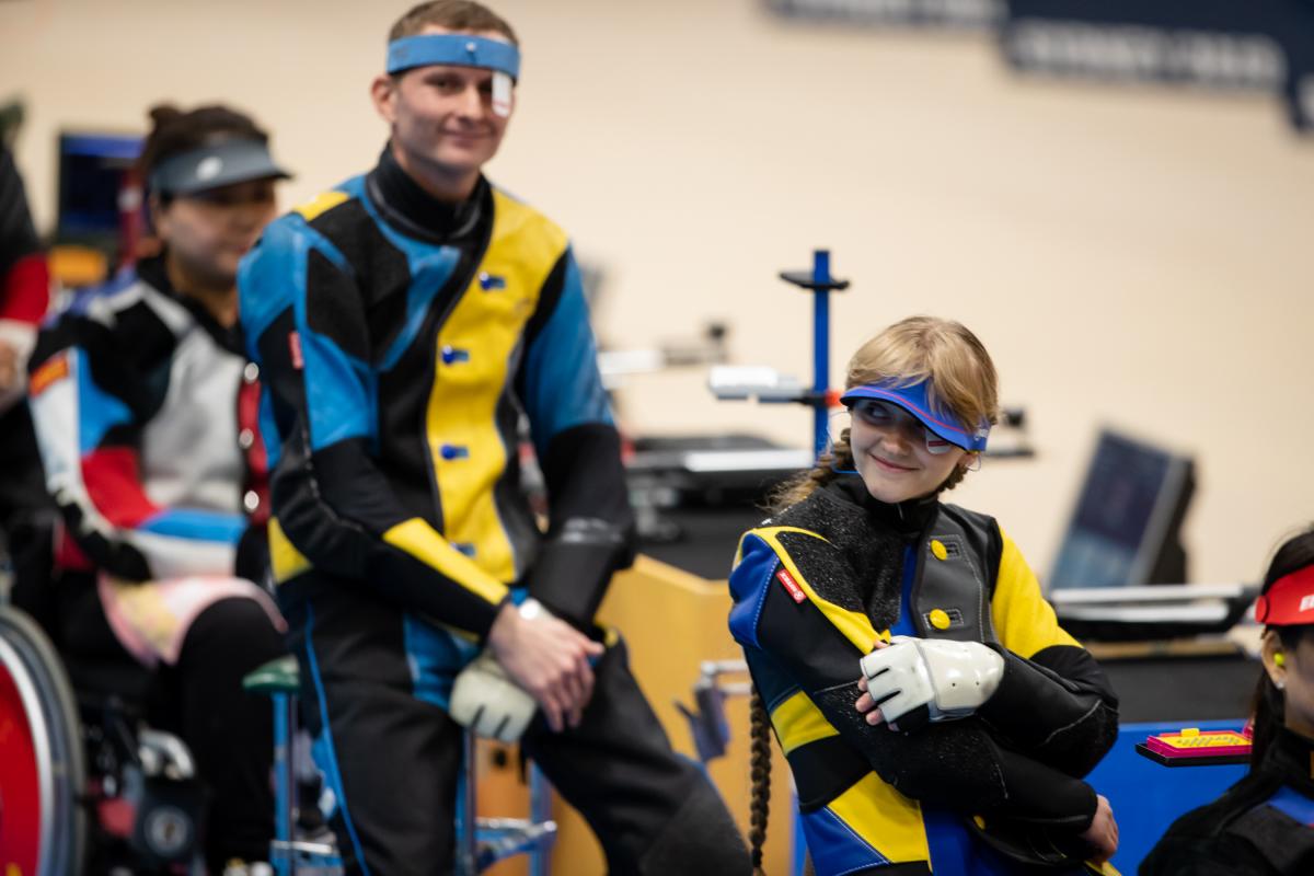 Ukrainian female shooter sits down smiling with male teammate behind also smiling