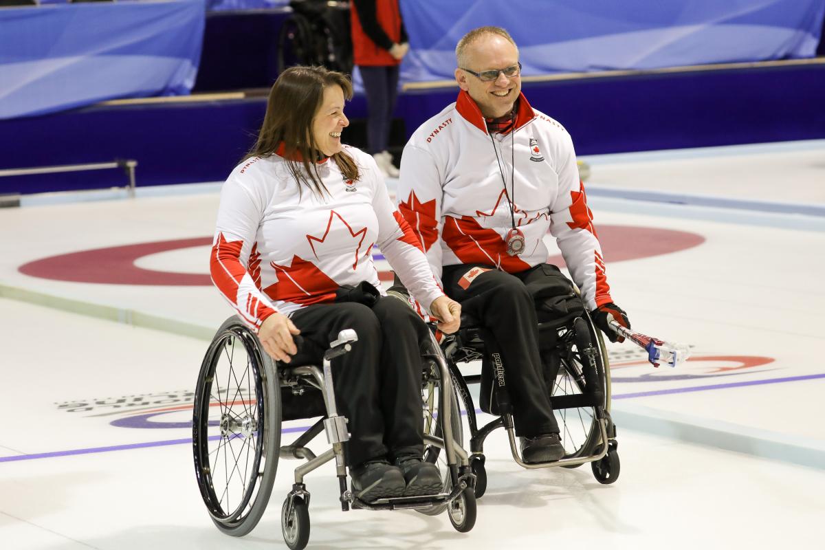 Man and woman in wheelchairs smile on curling ice sheet