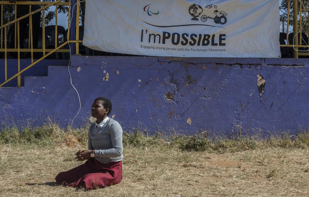 Student sits on the grass during an I'mPOSSIBLE activity in Malawi, with a flag of the programme in the background