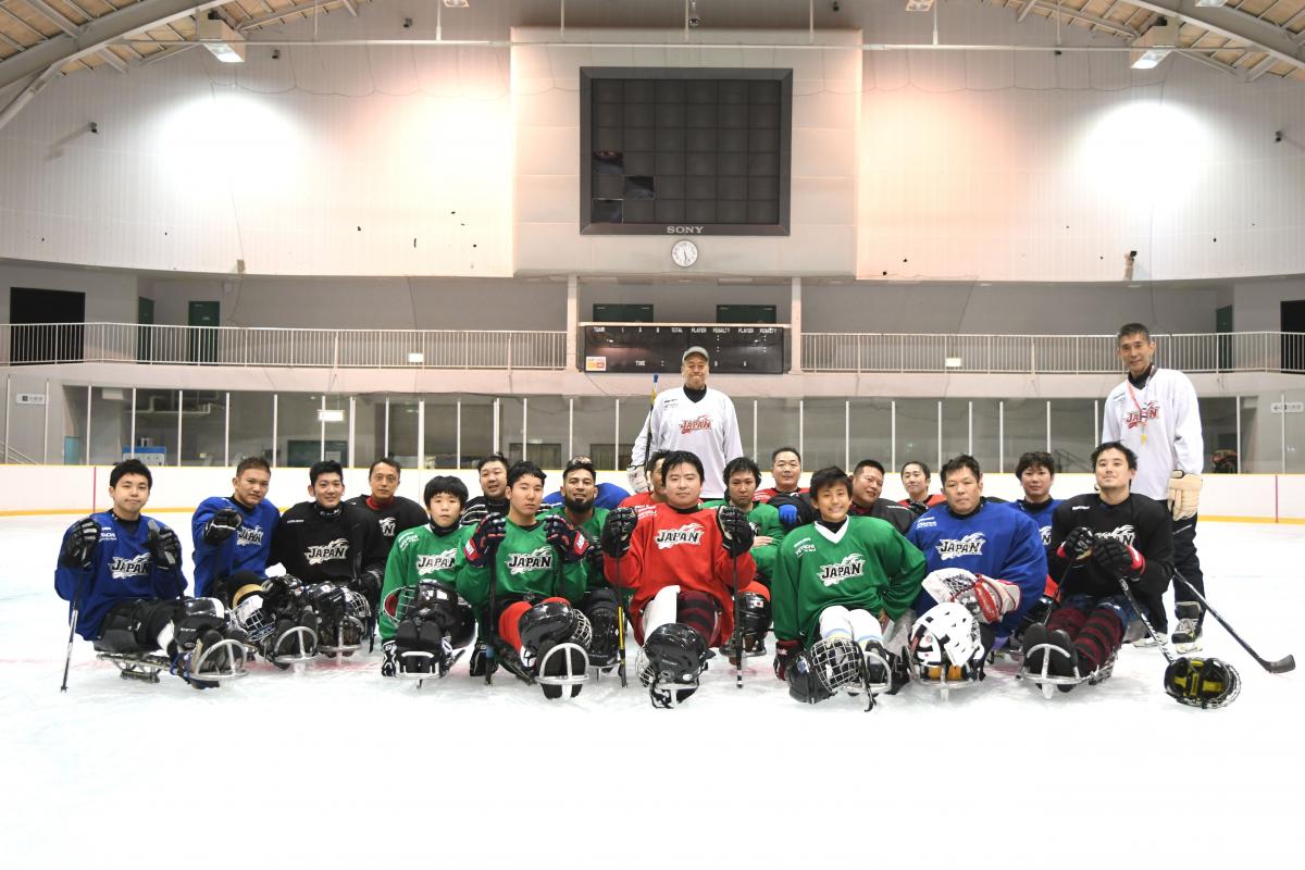 Two men standing on a ice rink behind a group of 20 Para ice hockey players on sledges