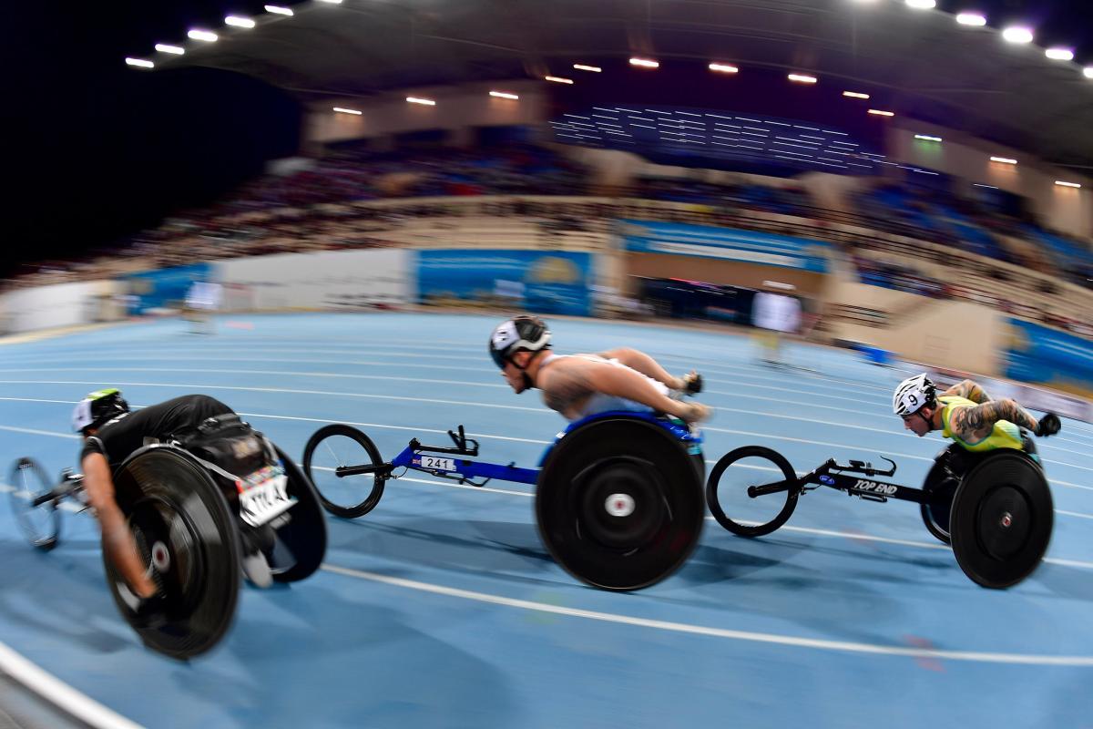 Three men in racing wheelchairs competing in a blue track