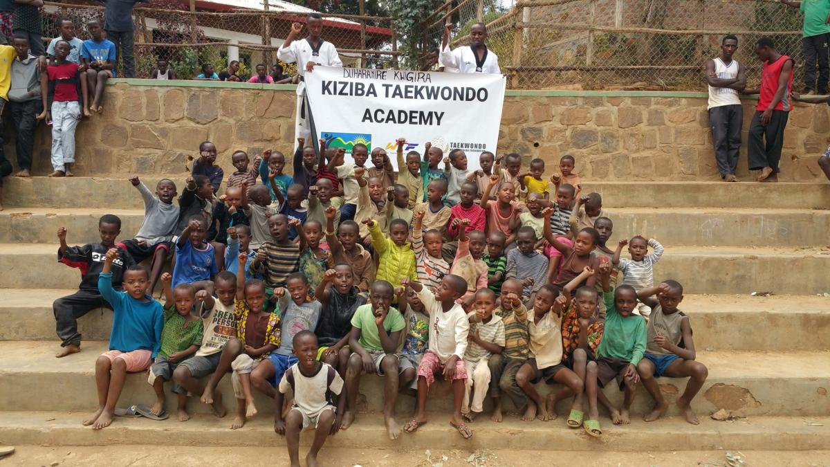 Group of African kids posing with fists raised and a banner behind saying Kiziba Taekwondo Academy