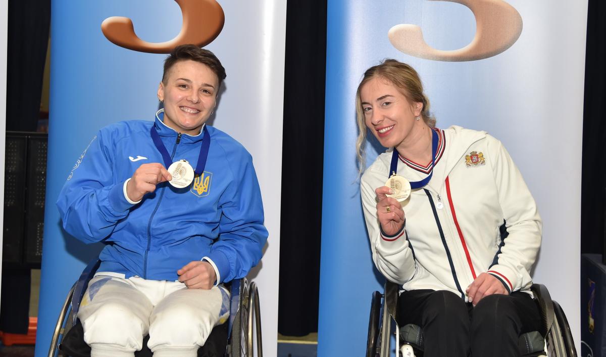 Two female wheelchair fencers pose with their medals on the podium