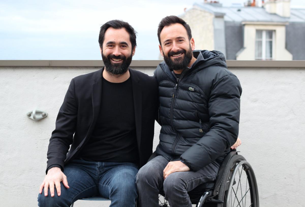 Two French males, one on a wheelchair, take a photo together smiling