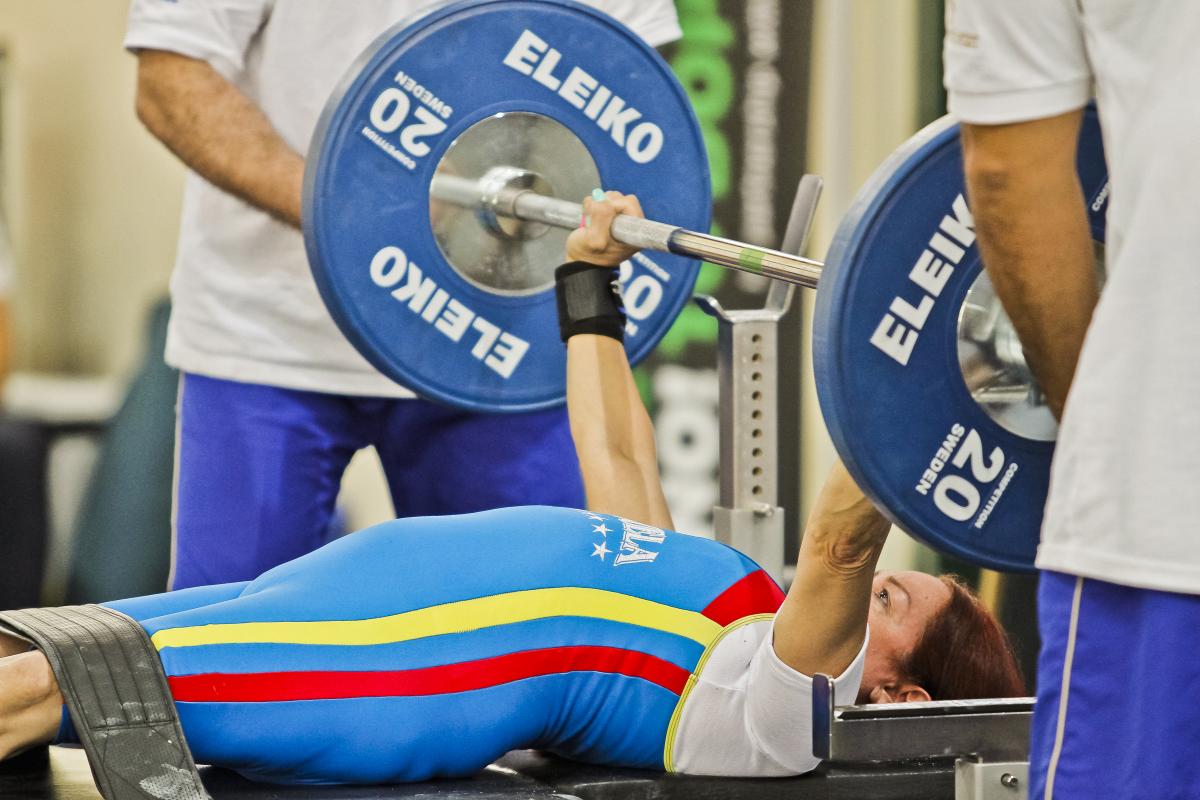 A woman lifting weight on a bench press with two standing men helping her