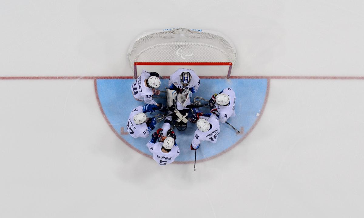 A group of six Para ice hockey players  huddle together in front of the goal