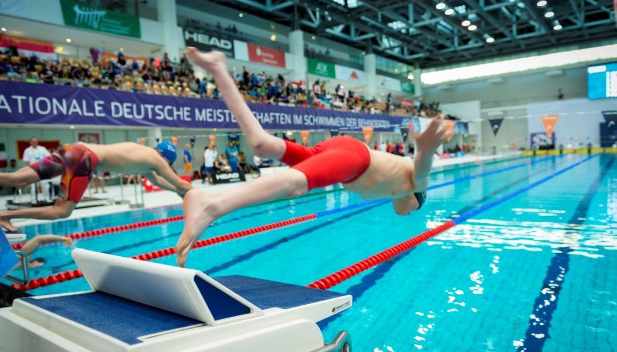 Two men jumping in the pool from starting blocks with people watching them from the stands