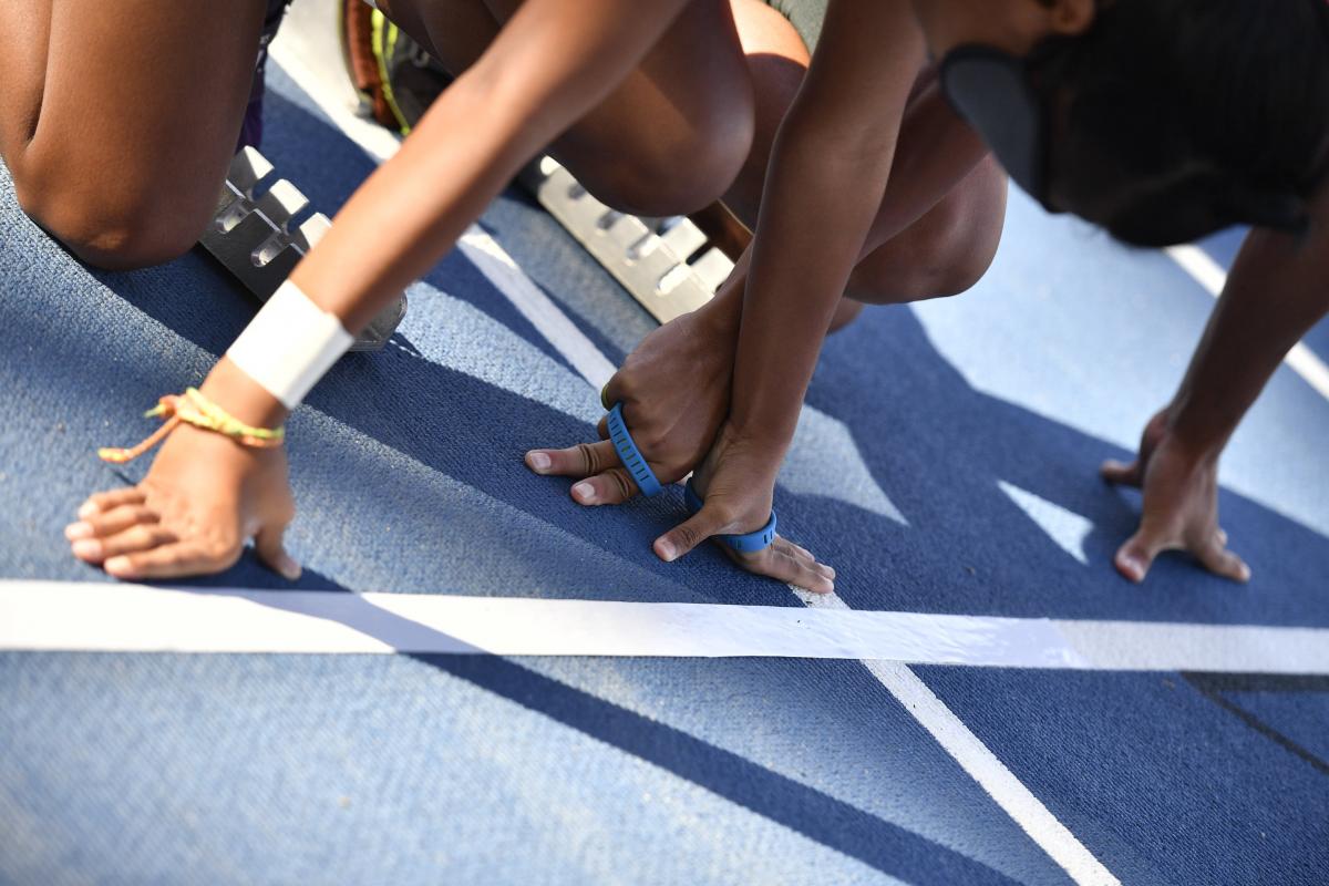 A blue athletics track with four hands side by side on the starting line