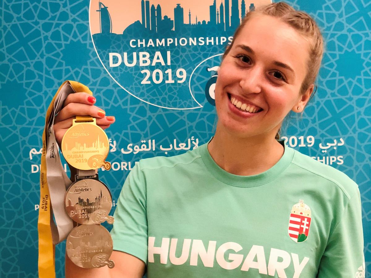 A young woman holding three medals and smiling with a shirt written Hungary