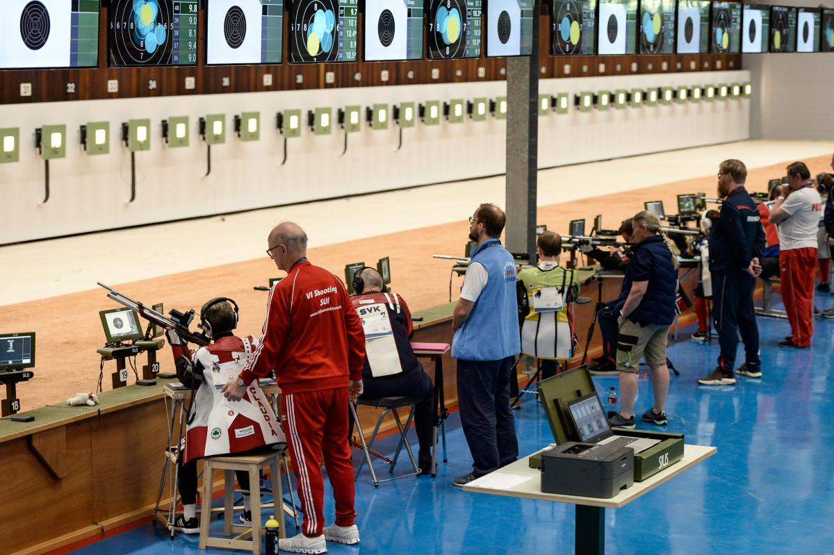 An indoor shooting range with five shooters with one person standing behind each of them
