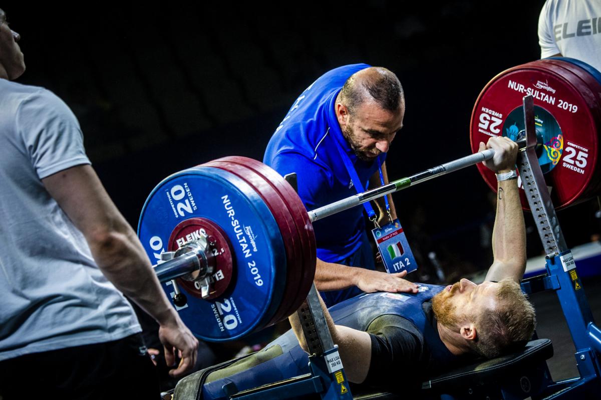 A man on a bench press preparing to lift a bar with three man standing around him