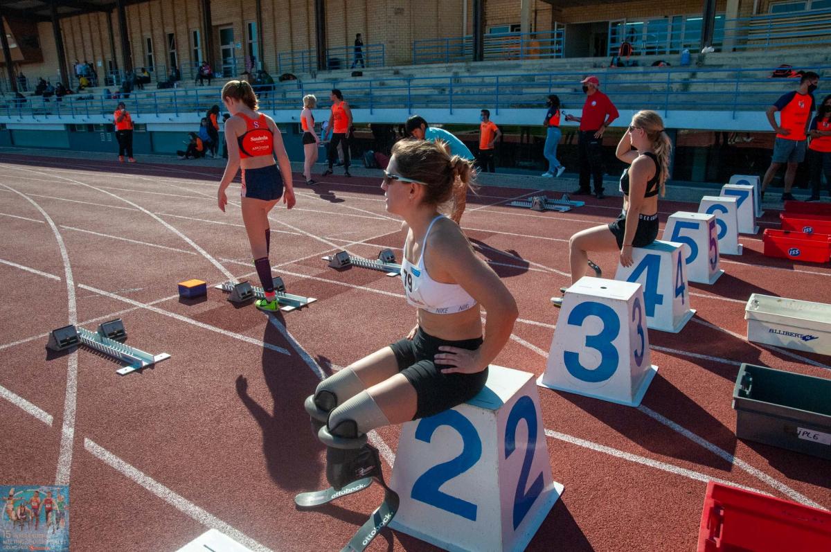Two women with prosthetic legs sitting on starting blocks on a red athletics track with two other women standing in front of them