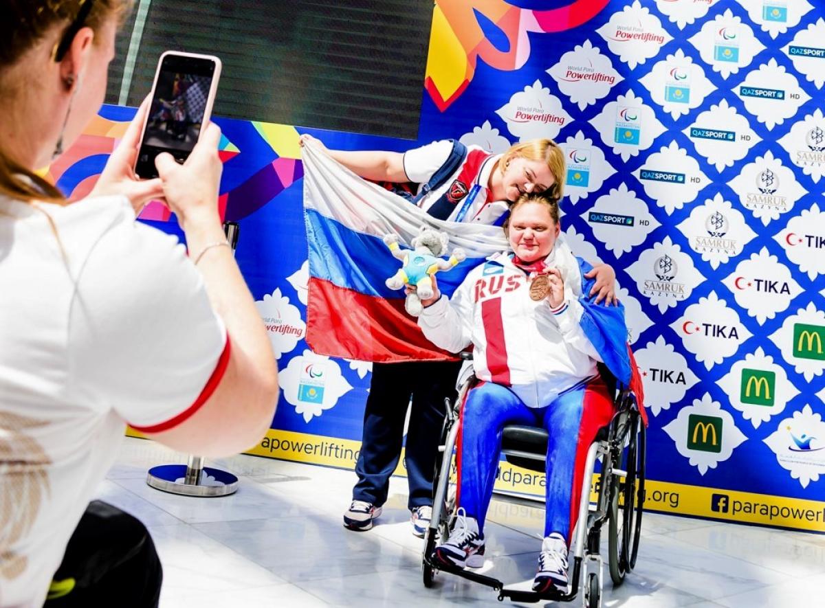 A woman in a wheelchair showing a medal and hugging another woman posing for a photo with the flag of Russia