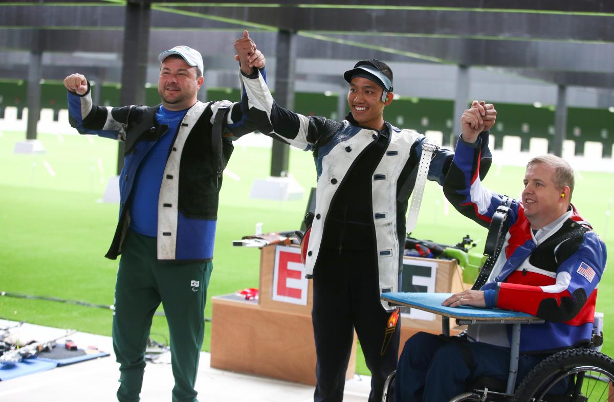 Two men standing and holding hands with a third man in a wheelchair in a shooting range