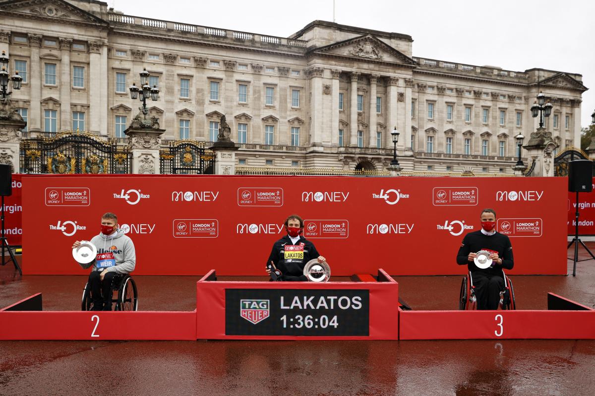 Three men in wheelchairs and face masks in a podium in front of Buckingham Palace in London