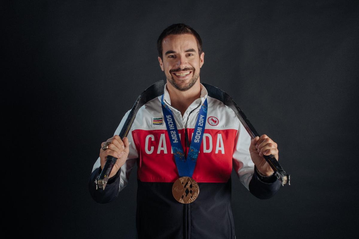 A smiling man with a bronze medal and wearing a Canadian jersey holds hockey sticks behind his neck