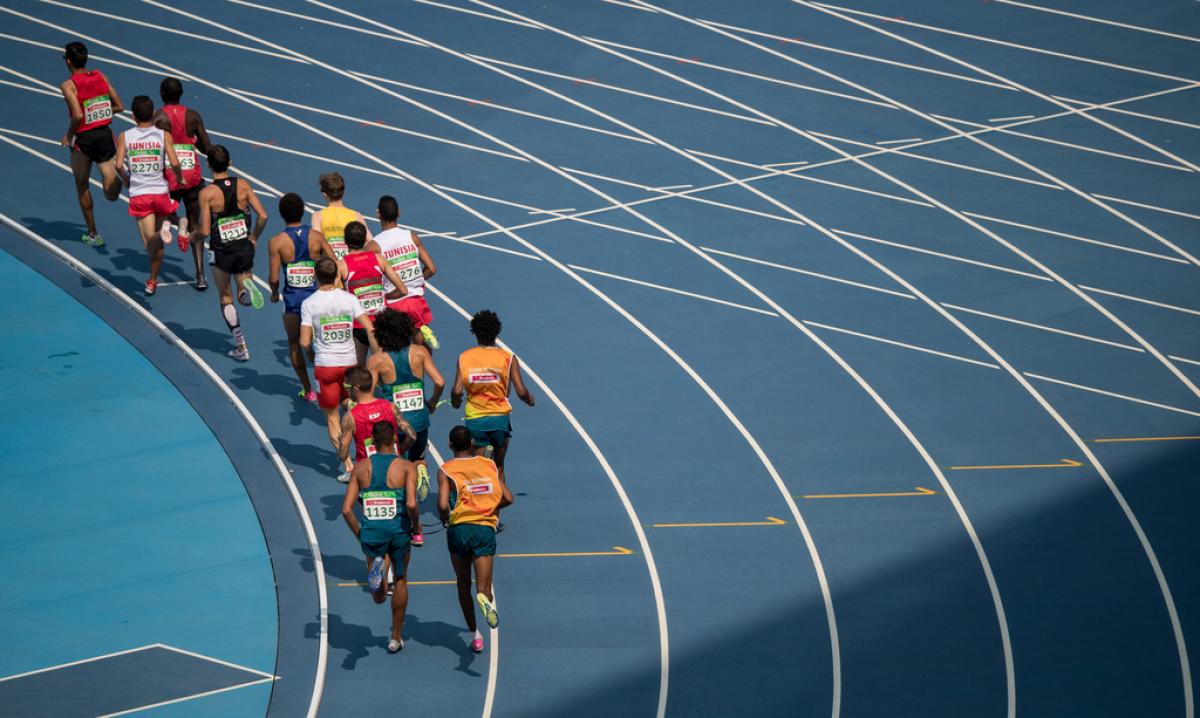 A group of 14 men seen from behind running in a blue athletics track