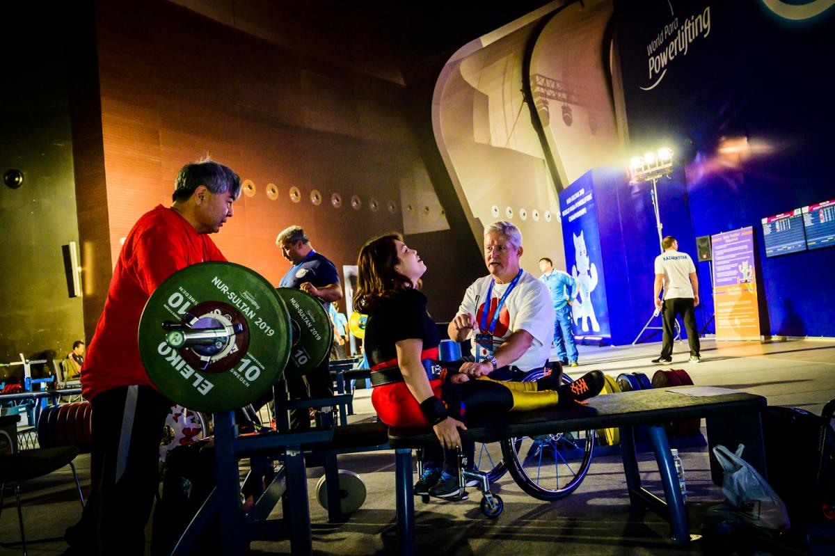 A woman on a bench press talking to a man in a wheelchair right next to her while another man stands near the barbell