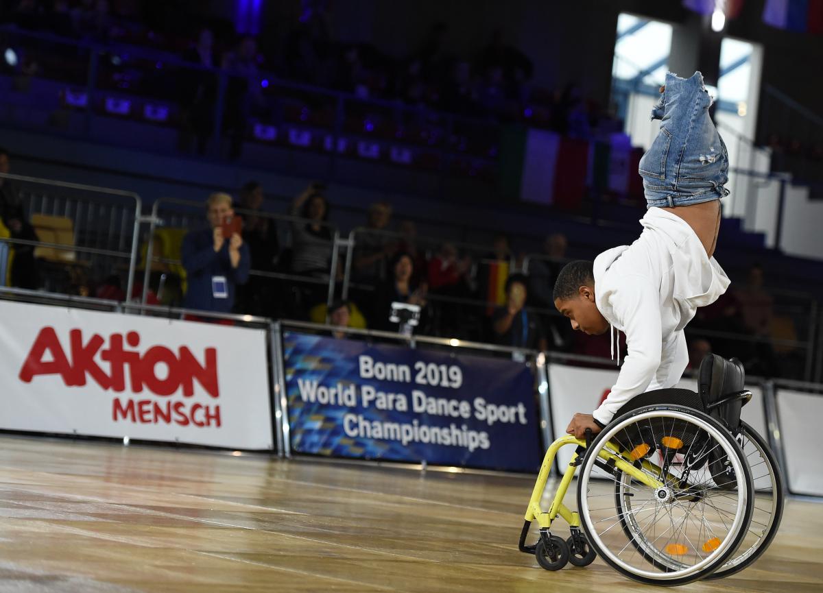 A man without legs upside down holding a wheelchair on a dancefloor 