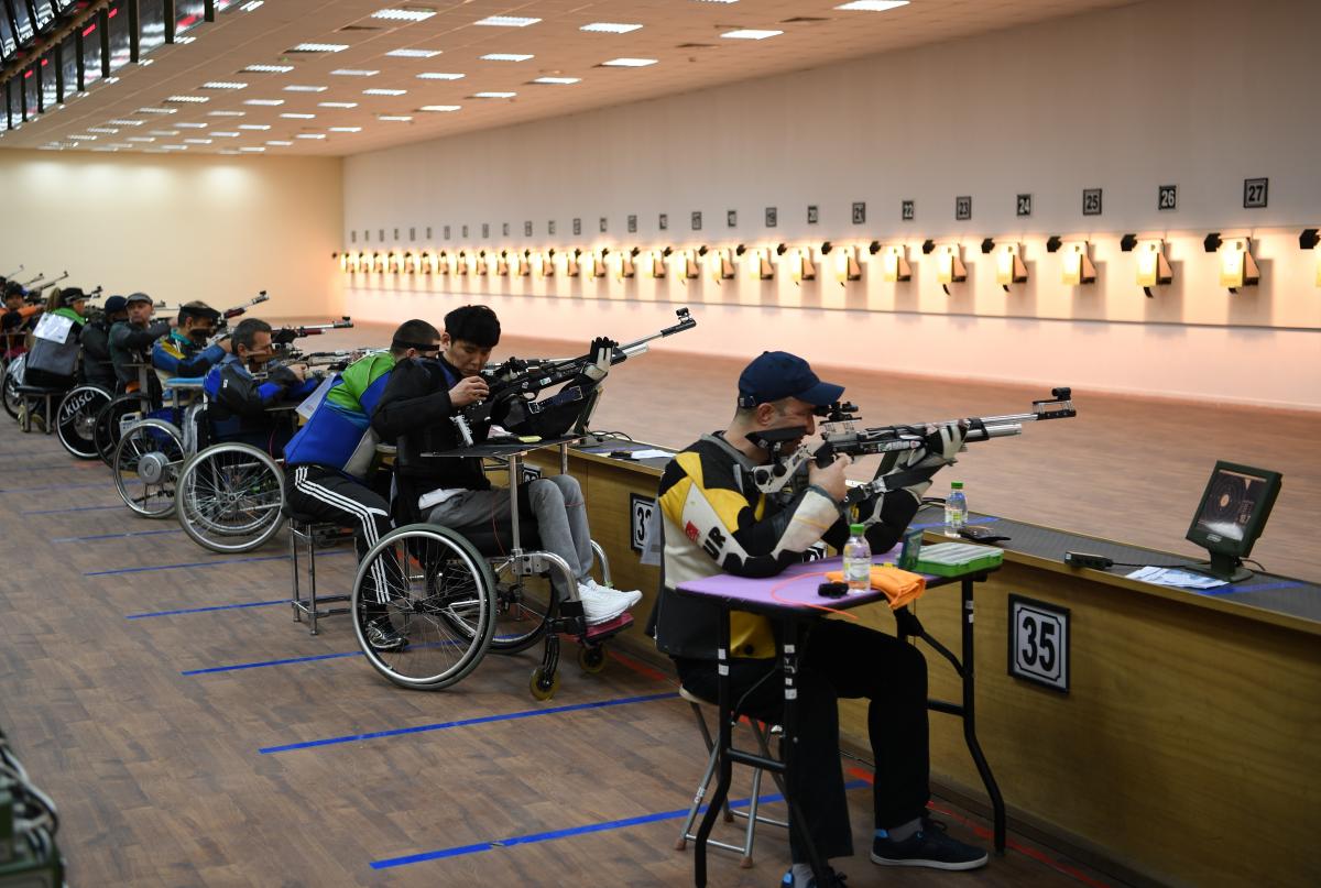 A group of men seated with rifles competing in an indoor shooting range