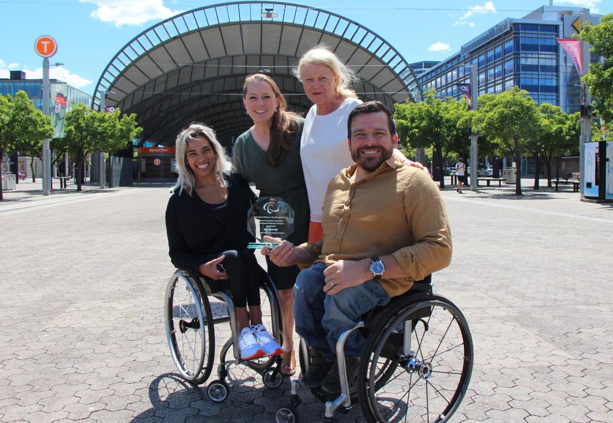 Two people in a wheelchair and two women standing smile with an award
