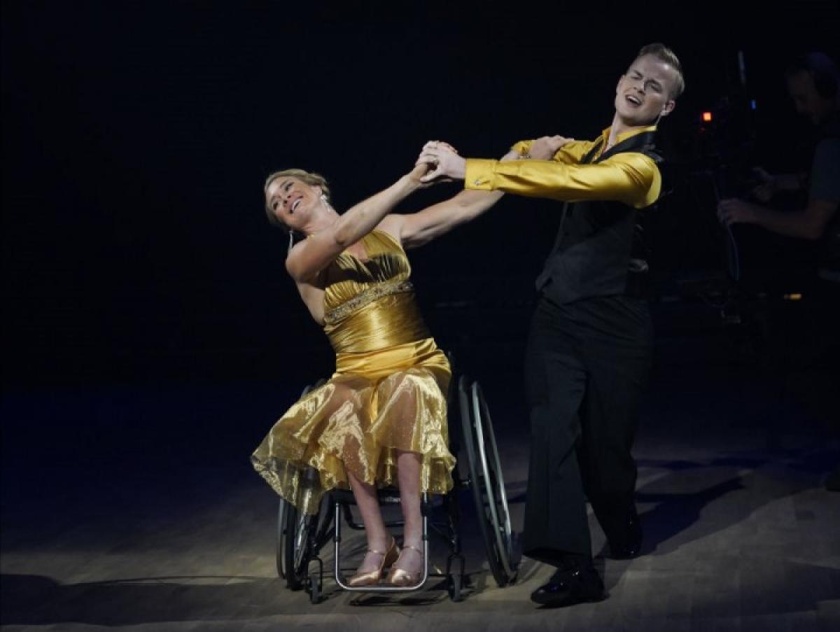 A female wheelchair dancer holding hands with her male standing dance partner on a dancefloor