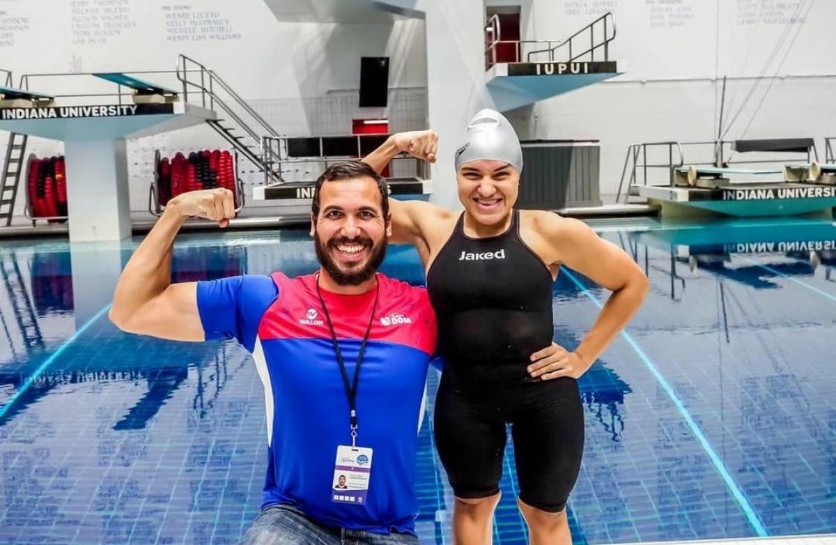 A short stature woman in swimming outfit with her arm around a knelled man with a swimming pool in the background