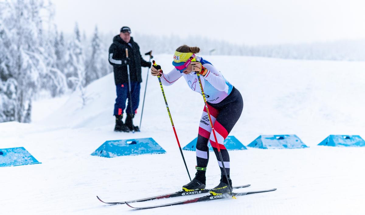 A female cross-country skier competing in the snow observed by a man in the background