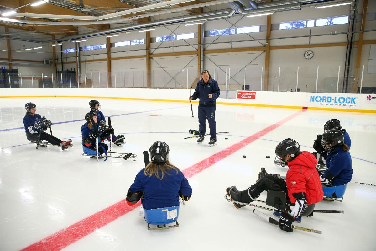 A woman on an ice rink talking to a group of seven people on Para ice hockey sledges 
