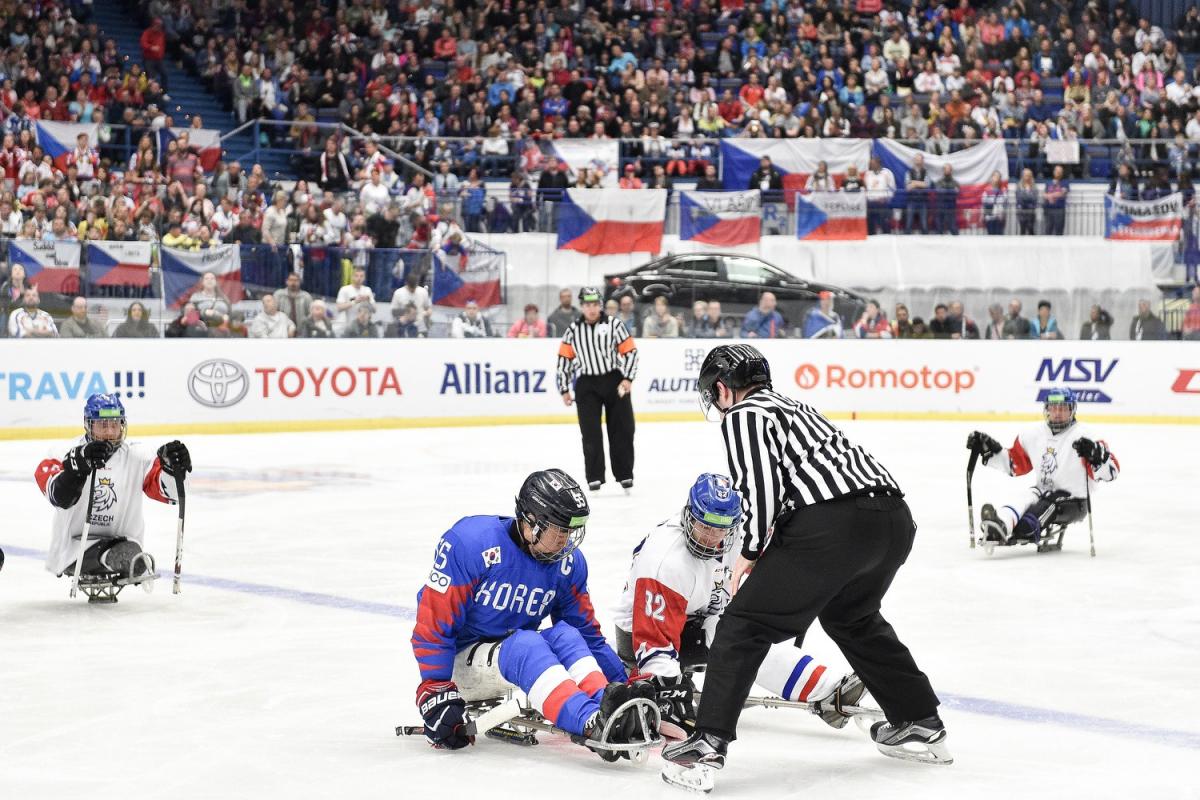 A referee throwing the puck in front of two players to start a Para ice hockey game in a crowded arena
