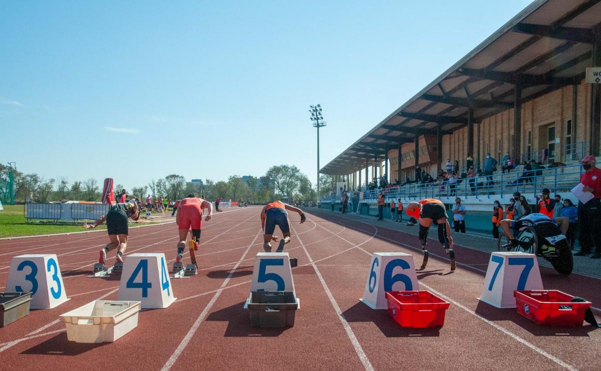 A red athletics track with athletes seeing from behind running from the starting blocks