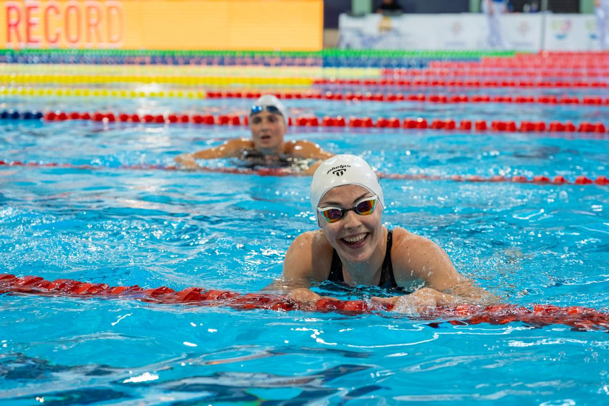 A young female swimmer with a white cap smiling in a swimming pool with another woman in the background