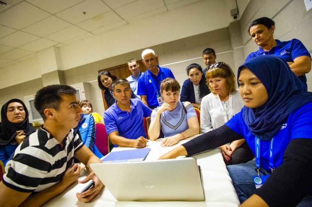 A group of 12 people around a table looking at a laptop