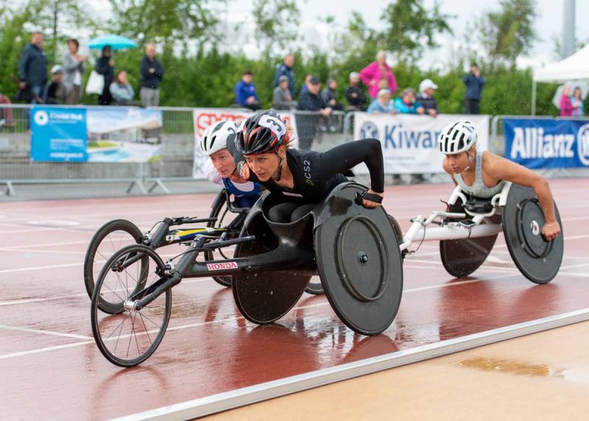 Three female wheelchair racers competing on a track under rain