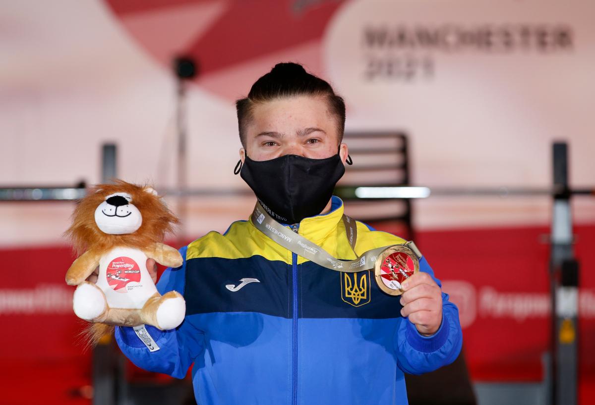 A woman with a face mask showing a medal and a Teddy bear mascot
