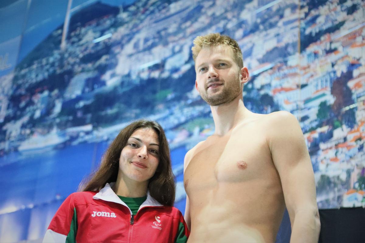 A short woman with a Portugal jumper and a tall shirtless man side by side in front of a panel in a swimming pool