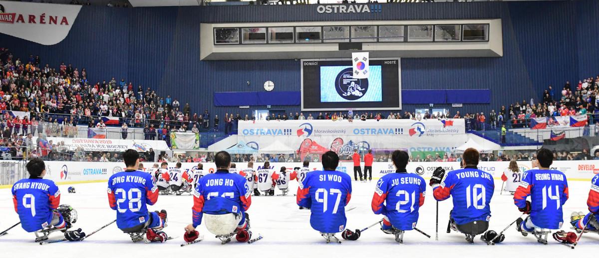 Two Para ice hockey teams on ice looking at a giant screen in a packed arena