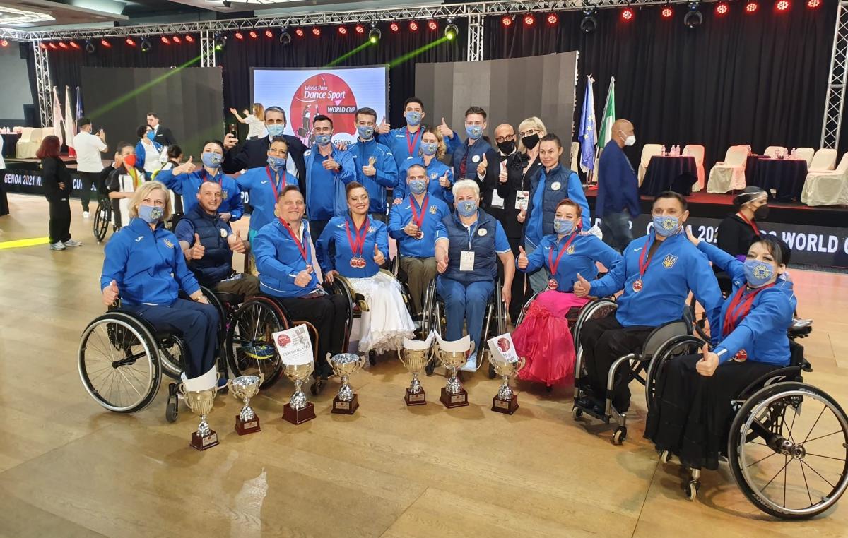 A group of nine people in wheelchairs with 12 people standing behind them posing for a picture with trophies in front of them