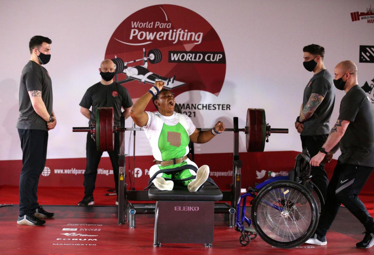 A woman celebrating on a bench press during a Para powerlifting event with four male loaders standing near her