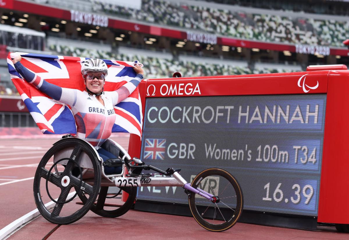 A woman in a racing wheelchair holding a British flag next to a board with her name and world record
